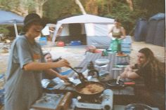 two people are cooking food on an outdoor stove in front of tents and picnic tables