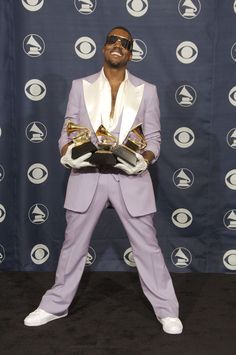 a man in a purple suit and white shoes holding two gold trophies while standing next to a black backdrop