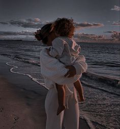 a woman holding a child on the beach at sunset with clouds in the sky behind her