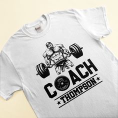 a white t - shirt with a man lifting a barbell and the words coach on it