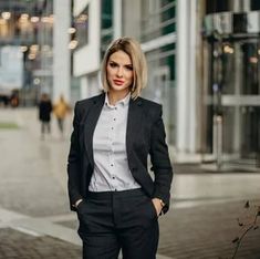 a woman standing on the sidewalk in front of a building wearing a black suit and white shirt