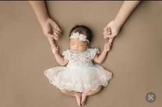 a baby is being held up by two hands while wearing a white lace dress and holding her mother's hand