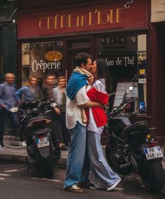 a man and woman kissing on the street in front of a cafe with people walking by