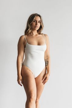 A woman standing in front of a white wall wearing a white one piece swimsuit with white spaghetti straps. White Fitted Swimwear With Low Back, Elegant Fitted Cream Swimwear, Elegant White Swimwear With Adjustable Straps, White Swimwear With Built-in Bra And Second-skin Fit, White Backless Swimwear With Straps, White Swimwear With Adjustable Strappy Back, White Strappy Back Swimwear With Adjustable Straps, White Swimwear With Adjustable Straps And Strappy Back, White Fitted Swimwear With Moderate Back Coverage