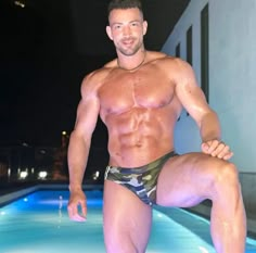 a shirtless man standing in front of a swimming pool at night with his legs spread out