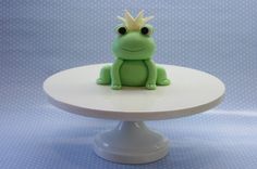 a green frog with a crown sitting on top of a white cake plate in front of a blue background