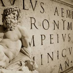 a statue of a man sitting on the side of a wall with roman writing behind it