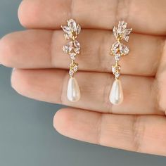 a pair of gold and white earrings with crystal leaves on the top, in front of a hand