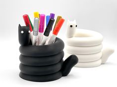 a black cat pen holder with markers and pens in it next to a stack of white cups