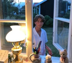 a young man standing in front of a window next to a white cat and lamp