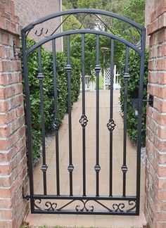 an iron gate in the middle of a brick walkway with green bushes and trees behind it