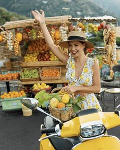 a woman riding on the back of a yellow scooter next to a fruit stand