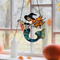 a stained glass mermaid hanging on a window sill in front of an orange pumpkin