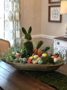 a wooden table topped with an easter basket filled with eggs and grass next to a window