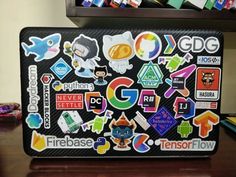 a laptop covered in stickers sitting on top of a wooden desk next to a book shelf