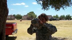 a woman in camouflage holding something up to her face while standing next to a tree