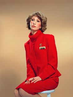 a woman in a red suit sitting on a stool with her legs crossed and looking off to the side