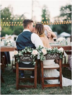 a bride and groom sitting at a wooden table with their names on it, surrounded by greenery