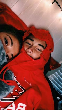 a man and woman laying in bed with red covers on their heads looking at the camera