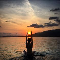 a person sitting in the water with their hands up at sunset or sunrise, while the sun is setting