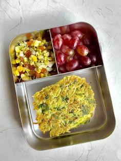 two metal trays filled with food on top of a white countertop next to grapes and corn