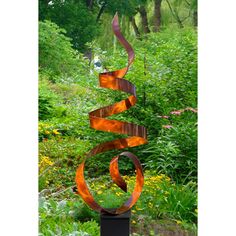 a tall metal sculpture in the middle of a garden with trees and bushes behind it