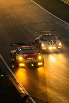 two racing cars driving down a race track in the rain at night with headlights on