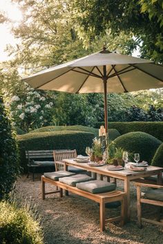 an outdoor dining table with chairs and umbrella in the middle of a garden area surrounded by hedges