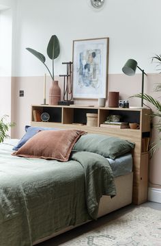 a bed with green sheets and pillows next to a book shelf filled with plants in front of a pink wall