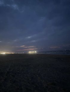 the sky is dark and cloudy at night, with lights shining in the distance on an empty beach