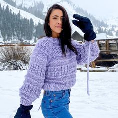 a woman is standing in the snow wearing a purple sweater and black gloves, with her hand up to her head