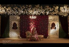 a bride sitting on a chair in front of a flower covered stage with flowers hanging from the ceiling