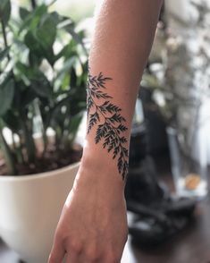 a woman's arm with a black leaf tattoo on her left wrist, and a plant in the background