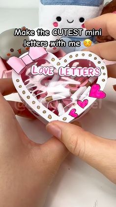 two hands holding a heart shaped box with letters inside it and a stuffed animal in the background