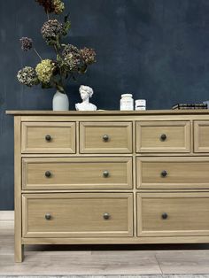 a wooden dresser with flowers on top and other items sitting on the bottom drawer, against a dark blue wall