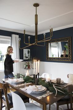 a woman is walking past a dining room table set with place settings, candles and greenery