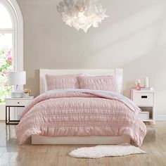 a bed with pink comforter and pillows in a white room next to a window