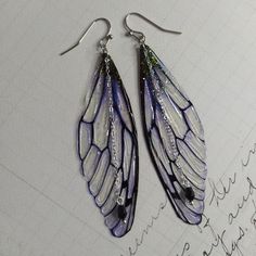 a pair of purple and white butterfly wings hanging from silver earwires on top of a piece of paper