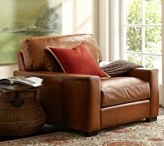 a brown leather chair sitting in front of a window next to a basket filled with books