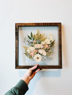 a person holding up a frame with flowers in it on the wall next to a white wall