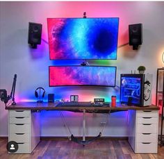 a computer desk with two monitors and speakers on top of it in front of a colorful wall