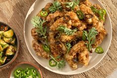 a white plate topped with fried food next to two bowls filled with green peppers and other vegetables