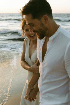 a man and woman walking on the beach holding each other's hands as they smile