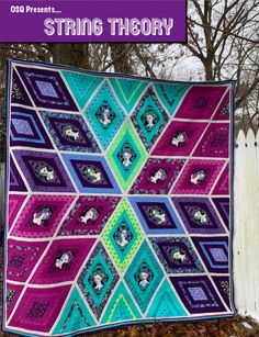 a colorful quilt hanging on the side of a white fence with trees in the background