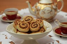 some cinnamon rolls on a white plate with tea in the back ground and two silver teapots