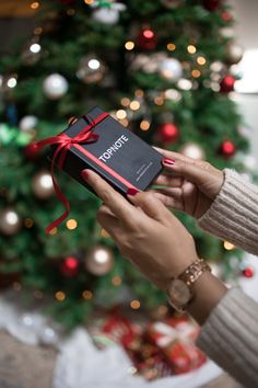 a woman holding a black box with a red ribbon around it in front of a christmas tree