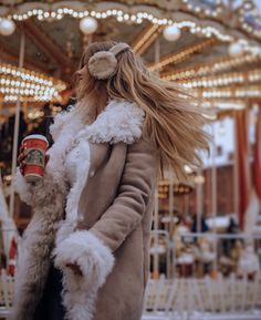 a woman in a fur coat is holding a coffee cup while standing near a merry go round