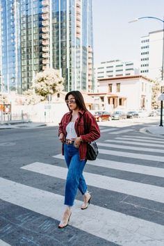 a woman walking across a cross walk in the middle of a city with tall buildings behind her