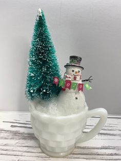 a snowman sitting in a cup with a small christmas tree