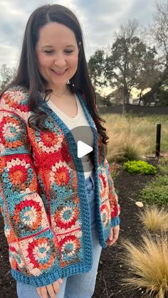 a woman standing in front of a bush wearing a colorful crocheted cardigan